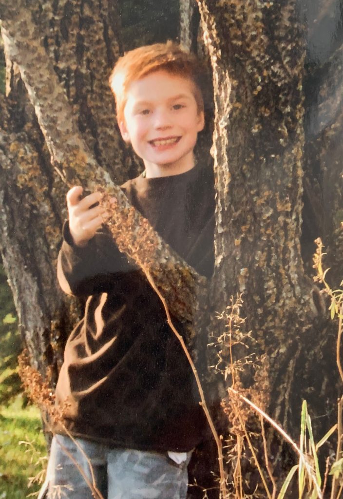 Luke Janzen at nine years old, a few months before his diagnosis, in a brown sweater, leaning against a tree and standing behind one of its branches